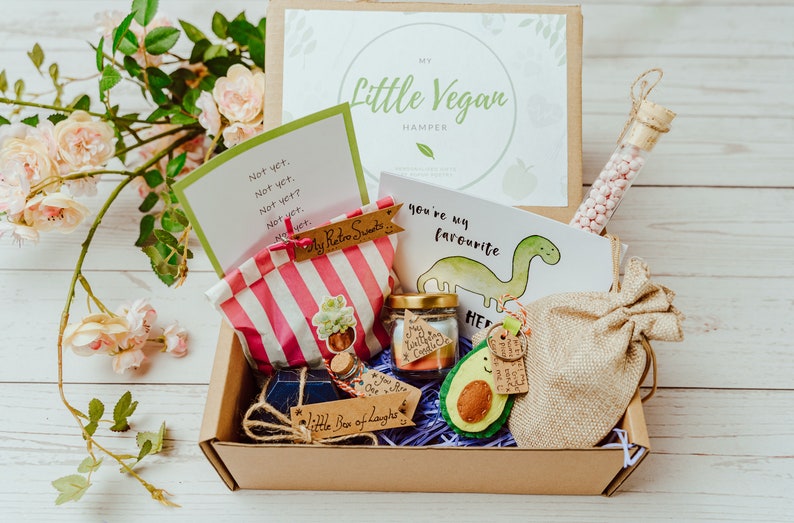 May include: A brown cardboard box filled with vegan treats, including a card that says "You're my favourite" with a dinosaur illustration, a bag of sweets, and a felt avocado. The box is labeled "My Little Vegan Hamper".