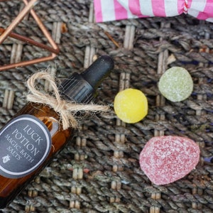 May include: A brown glass bottle labeled "LUCK POTION MAGIC BATH SALTS" with a dropper, tied with twine. Three colorful candies are scattered nearby. The items rest on a woven surface, with a copper-colored geometric structure in the background.