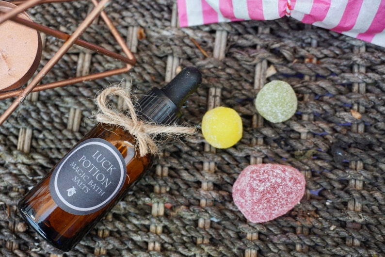 May include: A brown glass bottle labeled "LUCK POTION MAGIC BATH SALTS" with a dropper, tied with twine. Three colorful candies are scattered nearby. The items rest on a woven surface, with a copper-colored geometric structure in the background.