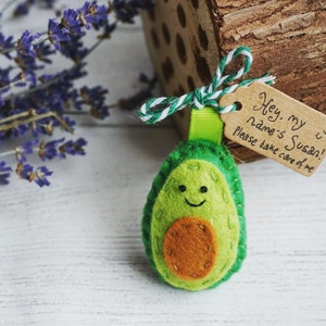 May include: A handmade felt avocado keychain with a smiling face and a brown seed. A small tag reads "Hey, my name's Susan! Please take care of me." The keychain is attached to a green ribbon and white and green twine.