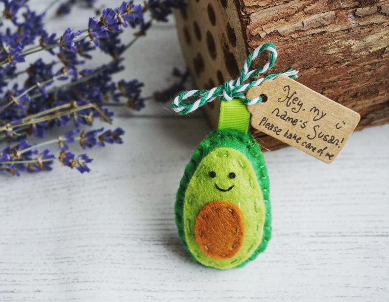 May include: A handmade felt avocado keychain with a smiling face and a brown seed. A small tag reads "Hey, my name's Susan! Please take care of me." The keychain is attached to a green ribbon and white and green twine.