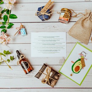 May include: An assortment of gifts and products arranged on a white wooden surface. Items include a small blue box, a candle in a jar, a burlap bag, a bottle of oil, a wrapped gift, a card with an avocado illustration, and a small bottle with blue candies.