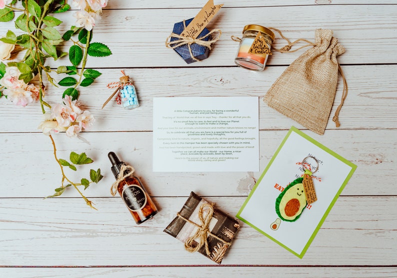 May include: An assortment of gifts and products arranged on a white wooden surface. Items include a small blue box, a candle in a jar, a burlap bag, a bottle of oil, a wrapped gift, a card with an avocado illustration, and a small bottle with blue candies.