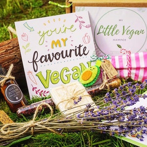 May include: A vegan gift set with a variety of items, including a card that says "You're my favourite Vegan", a bottle of bath salts, a bar of soap, a small jar of pink candies, and a bundle of lavender. The set is arranged on a bed of green grass.