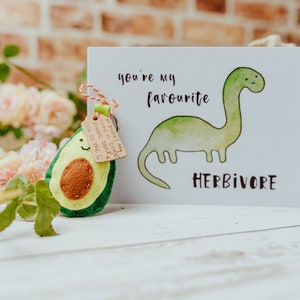 May include: A greeting card with a watercolor illustration of a green dinosaur and the words "you're my favourite herbivore". An avocado-shaped felt keychain is attached with a tag. The background includes flowers and a burlap bag.