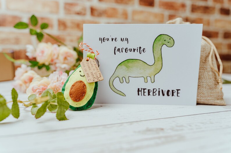 May include: A greeting card with a watercolor illustration of a green dinosaur and the words "you're my favourite herbivore". An avocado-shaped felt keychain is attached with a tag. The background includes flowers and a burlap bag.