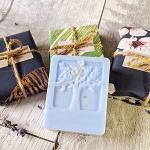 May include: A light blue soap bar with a tree design, next to three wrapped packages tied with twine. The soap has the word "Flower" on it. The packages are wrapped in patterned paper.