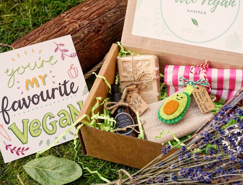 May include: A vegan gift hamper with a greeting card that reads "You're my favourite Vegan." The box contains a bar of soap, a bottle of "Luck Potion" bath oil, a wrapped gift, and an avocado-shaped felt ornament. The box is filled with green paper shreds.