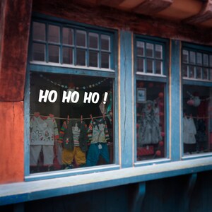 Puede incluir: Un escaparate de tienda con el texto "Ho Ho Ho!" en letras blancas. El escaparate est&aacute; decorado con luces de Navidad y muestra ropa para ni&ntilde;os en varios colores y dise&ntilde;os.