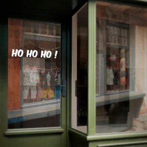 Puede incluir: Una fachada de tienda verde con una puerta de vidrio. La puerta tiene un letrero que dice "HO HO HO!" en letras blancas. La fachada est&aacute; decorada con luces de Navidad y un &aacute;rbol de Navidad.