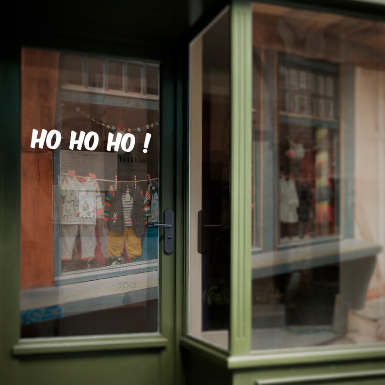 Puede incluir: Una fachada de tienda verde con una puerta de vidrio. La puerta tiene un letrero que dice "HO HO HO!" en letras blancas. La fachada est&aacute; decorada con luces de Navidad y un &aacute;rbol de Navidad.