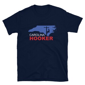 Pode incluir: Camiseta azul marinho com um contorno azul da Carolina do Norte e o texto "CAROLINA HOOKER" em vermelho e branco. O design inclui uma silhueta de uma pessoa em um poste de eletricidade.