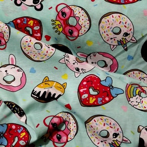 May include: A light blue fabric with a repeating pattern of cartoon donuts. The donuts have various designs, including sprinkles, crowns, and animal faces like cats, bunnies, and pandas. Some donuts are heart-shaped with the word "LOVE".