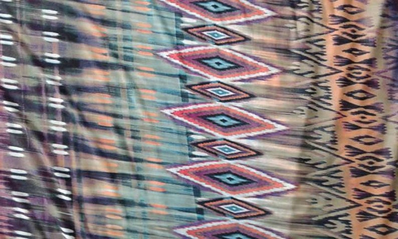 May include: A fabric with a colorful geometric pattern. The pattern features a repeating diamond shape with shades of orange, blue, purple, and green. The fabric has a soft, flowing texture.