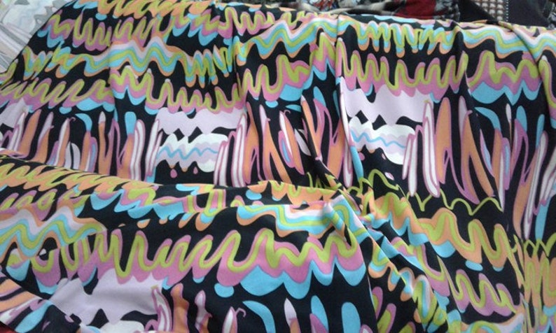 May include: A colourful fabric with an abstract design. The pattern includes wavy lines and shapes in black, pink, blue, orange, and yellow. The fabric is draped, highlighting the pattern's flow and texture. Ideal for clothing or home decor.