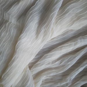 May include: A close-up of a white sheer fabric with a crinkled texture. The fabric is draped and folded, creating soft, flowing lines.