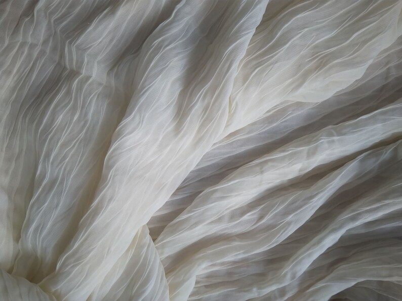 May include: A close-up of a white sheer fabric with a crinkled texture. The fabric is draped and folded, creating soft, flowing lines.
