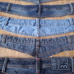 Könnte beinhalten: Fünf verschiedene Jeans-Bundbänder, aus Jeans geschnitten, auf einer Holzoberfläche ausgelegt. Die Bundbänder sind alle in verschiedenen Blautönen gehalten und haben unterschiedliche Nähdetails. Das obere Bundband hat ein braunes Leder-Etikett mit dem Text "NEXT DENIM" darauf. Das untere Bundband hat ein braunes Leder-Etikett mit dem Text "CLINK JEANS LONDON" und der Nummer "33" darauf.