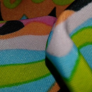 May include: Close-up of a vibrant fabric with a retro design. The fabric displays a pattern of curved lines and shapes in shades of blue, green, orange, pink, and black. The fabric is folded, revealing the texture and detail of the material.