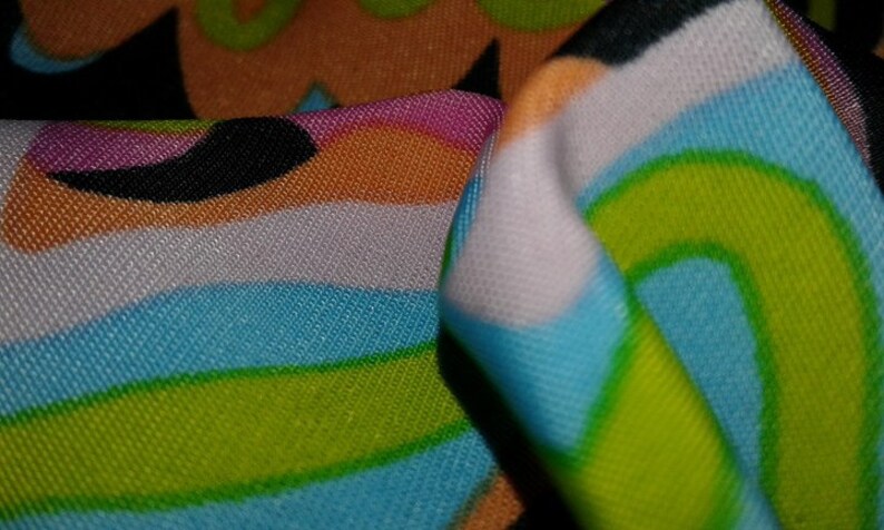 May include: Close-up of a vibrant fabric with a retro design. The fabric displays a pattern of curved lines and shapes in shades of blue, green, orange, pink, and black. The fabric is folded, revealing the texture and detail of the material.