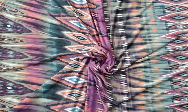 May include: A close-up of a fabric with a colorful ikat pattern. The fabric is a blend of purple, orange, green, and black. The pattern is a repeating diamond shape with a white outline.