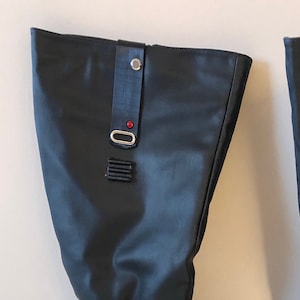 May include: A pair of black knee-high boots. The boots have a faux leather appearance with a small heel. They feature a decorative strap with a silver buckle and a small red accent near the top. White stitching accents the ankle area.