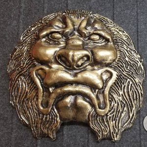 Puede incluir: Dos esculturas murales de caras de león de color bronce. La escultura más grande mide aproximadamente 25 cm de ancho y la más pequeña, unos 13 cm. Ambas tienen melenas y rasgos faciales detallados, con una moneda para comparar el tamaño.