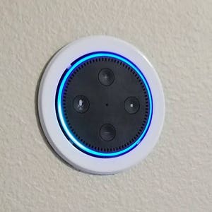 Peut inclure: Un haut-parleur intelligent rond blanc avec un visage noir et un anneau lumineux bleu autour du bord. Le haut-parleur est monté sur un plafond blanc.