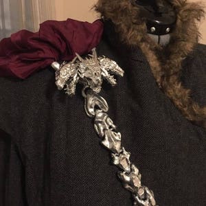 Könnte beinhalten: Dekorative silberfarbene Brosche mit Drachendesign. Die Brosche ist an einem dunkelgrauen Fischgrätenmantel befestigt, mit einem bordeauxfarbenen Stoff und einem braunen Pelzkragen.