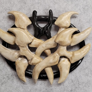 以下が含まれることがあります： スタイリッシュなデザインの装飾壁掛け。黒い円形のフレームに、外側に放射状に広がるオフホワイトの爪のような形が特徴です。上部には黒い、様式化された手の形があります。背景は明るい灰色の生地です。