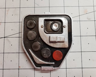 May include: A rectangular, metallic-looking device with a black and silver color scheme. It features several circular buttons, a central lens, and a red button. The device has a slightly worn appearance.