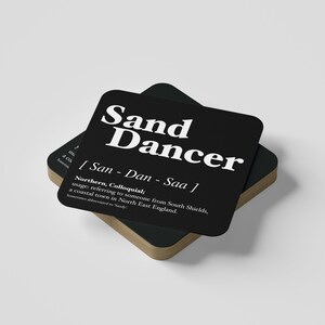 May include: Two square black coasters with the text "Sand Dancer" in white. The coasters have a definition below the title, with the words "Northern, Colloquial" and "South Shields" visible. The coasters have a light brown wooden edge.