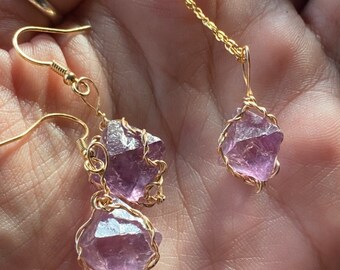 May include: A gold-colored necklace and earrings set featuring raw amethyst crystals. The crystals are wrapped in gold wire and suspended from delicate gold chains and earring hooks. The amethyst has a light purple hue.