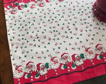 Op de afbeelding: Een rechthoekige kersttafelloper met een herhalend patroon van kerstmannengezichten langs de rode randen. Het midden heeft een witte achtergrond met kleine groene hulst- en rode bessenontwerpen. Een feestelijke vakantieversiering.
