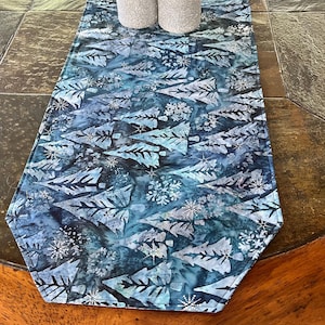 May include: A blue table runner with a white snowflake and tree pattern. The runner has a scalloped edge and is made of fabric.