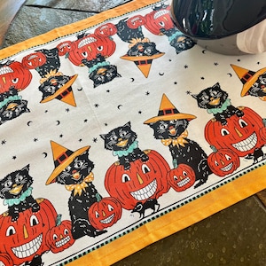 May include: A white fabric table runner with a black and orange Halloween theme. The runner features black cats wearing witch hats and sitting on jack-o'-lanterns. The runner has a yellow border and a white background with black stars and crescent moons.