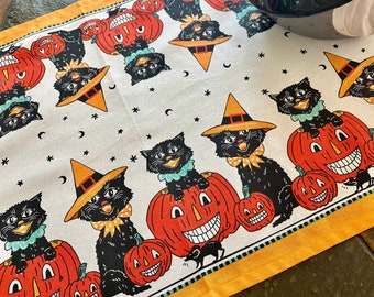 May include: A white fabric table runner with a black and orange Halloween theme. The runner features black cats wearing witch hats and sitting on jack-o'-lanterns. The runner has a yellow border and a white background with black stars and crescent moons.