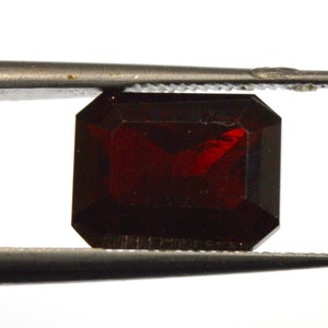 May include: A deep red gemstone, likely a garnet, is held by tweezers over a ruler marked with numbers 10 and 11. The gemstone is a rectangular emerald cut.