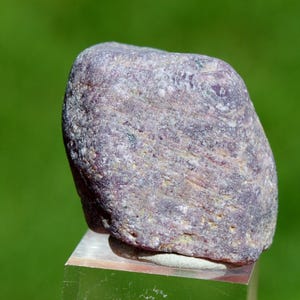 Könnte beinhalten: Ein rauer, abgerundeter Stein mit einem gesprenkelten Aussehen, der Farbtöne von Lila, Grau und Grüntönen aufweist. Der Stein ist vor einem verschwommenen grünen Hintergrund platziert, der seine natürliche Textur und Farbvariationen hervorhebt. Der Stein ruht auf einem transparenten, rechteckigen Ständer.