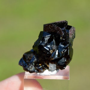 Puede incluir: Un grupo de cristales negros, lustrosos y facetados sobre un soporte de exhibición transparente. Los cristales tienen una forma geométrica aguda y una apariencia oscura, casi opaca.