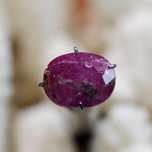 Pode incluir: Uma gema oval de cor rosa escura com corte facetado. A gema é mantida no lugar por quatro garras de metal.