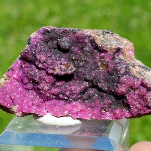 May include: A pink and black cobalt mineral specimen with a rough, textured surface. The mineral is on a clear display stand.