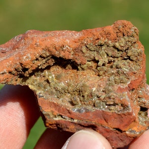 Puede incluir: Primer plano de una roca rugosa de color marrón rojizo con una cavidad llena de pequeños cristales verdes y blancos.