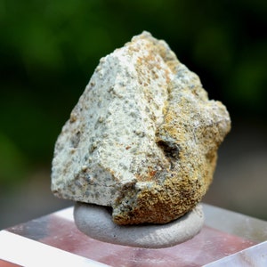 May include: A rough, grey and brown rock with a textured surface. The rock is sitting on a white display stand.