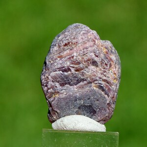 Könnte beinhalten: Ein natürlicher, grob geschnittener Edelstein mit geschichteter Struktur. Der Stein zeigt Schattierungen von Lila, Braun und Grau und ruht auf einem kleinen, weißen Sockel. Der verschwommene grüne Hintergrund hebt die Textur und Farbvariationen des Steins hervor.