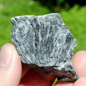 May include: A close-up of a dark gray rock with a striated texture. The rock has a rough, uneven surface and appears to be made of multiple layers.