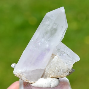 Peut inclure: Un groupe de cristaux de quartz transparents avec une touche de violet, posés sur une base blanche. Les cristaux sont pointus et ont une texture naturelle et rugueuse.