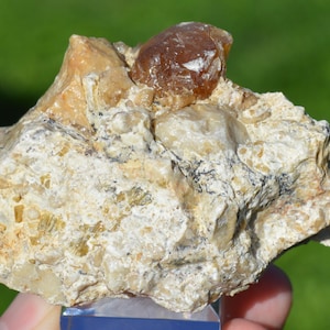 May include: A rough, textured rock formation with a light beige and off-white color palette. A dark brown, rounded stone sits atop the rock. The rock has a natural, organic appearance, with visible mineral deposits and variations in texture.