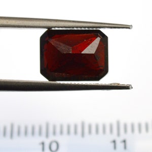 May include: A deep red gemstone with a rectangular cut, held in tweezers over a ruler marked in centimeters.