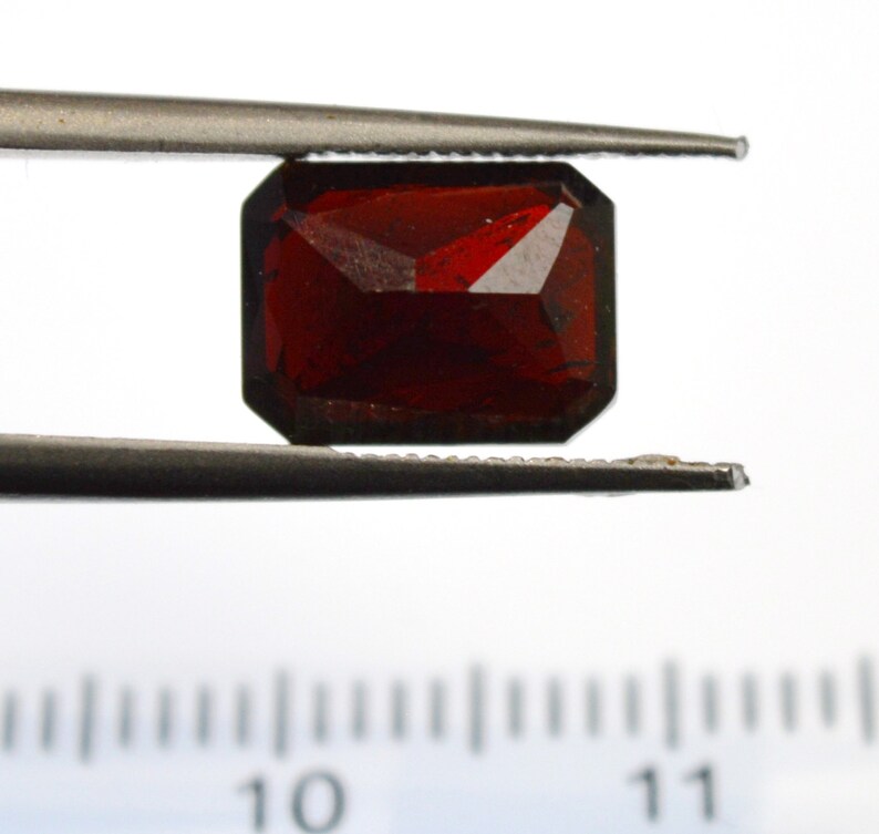 May include: A deep red gemstone with a rectangular cut, held in tweezers over a ruler marked in centimeters.